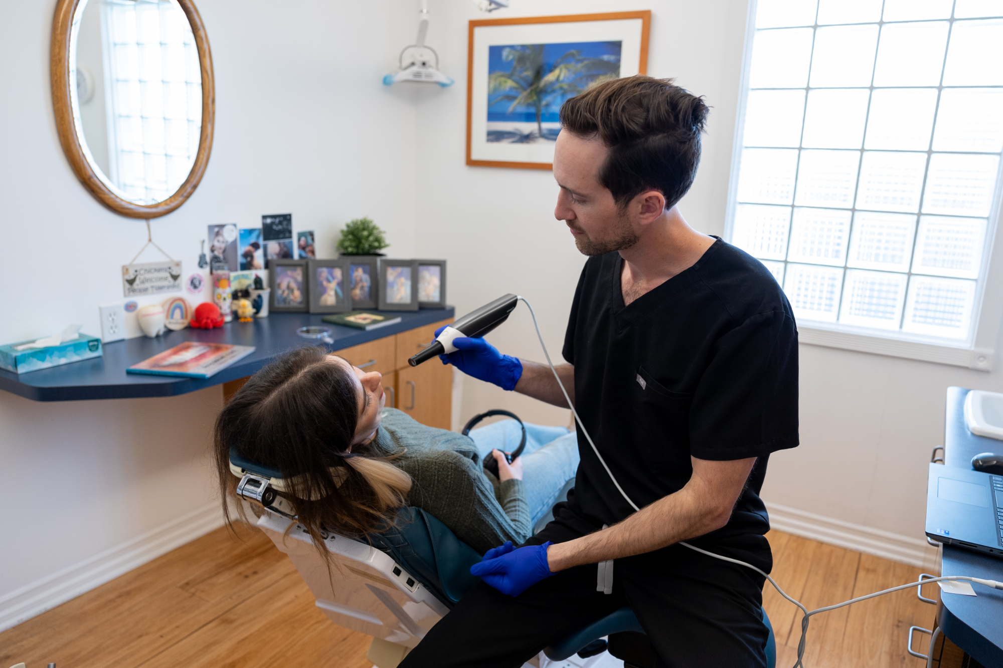 Patient with Dr. Keith during an appointment - Hillcrest Dental Dr. Keith, a man with short dark hair in a black scrub, holds a dental tool. A young woman is reclining in the dental chair, holding headphones - Hillcrest Dental