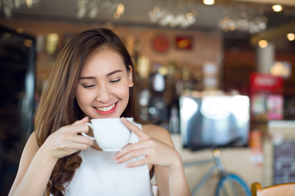 Smiling Woman with Coffee at Café – Clear Aligners A young woman with long hair smiles while looking down at a white cup in her hands, seated in a cozy café – Clear Aligners