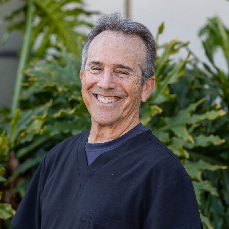 Dr. Doug Disraeli - Hillcrest Dental A headshot of Dr. Doug Disraeli, a smiling man with short gray hair, wearing a black medical scrub top, standing outdoors with greenery in the background. - Hillcrest Dental