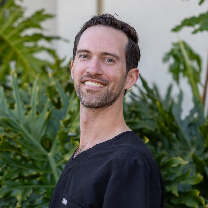 Dr. Andrew Keith - Hillcrest Dental A professional headshot of Dr. Andrew Keith, a smiling man with short dark hair, wearing a black shirt against a tropical leafy background. - Hillcrest Dental