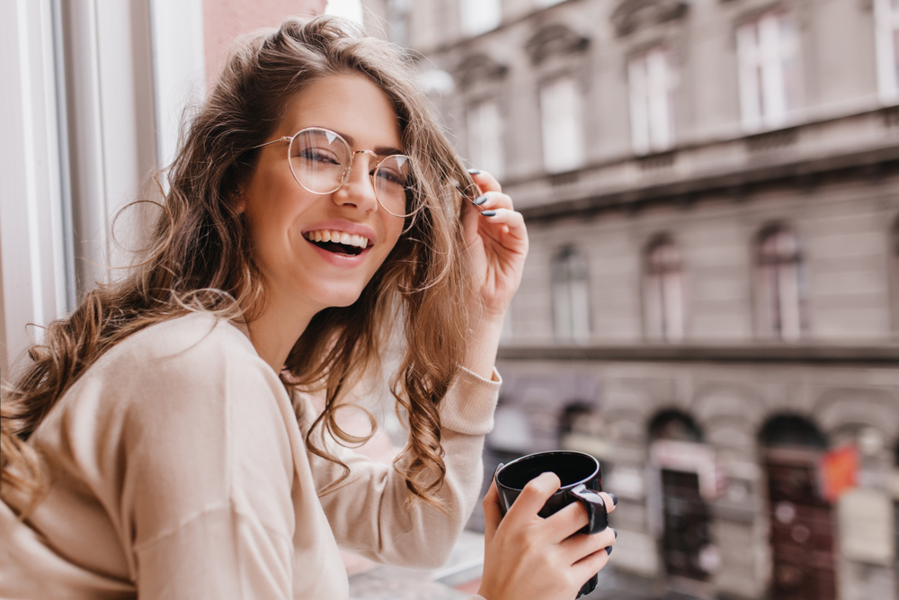 Smiling Woman with Glasses Holding Coffee – Clear Aligners A cheerful woman wearing glasses and holding a black coffee mug smiles brightly while sitting by a window in an urban setting – Clear Aligners